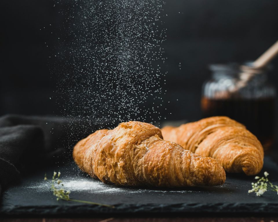 Two freshly baked croissants dusted with powdered sugar on a dark slate board, with a jar of preserves and a honey dipper behind them.