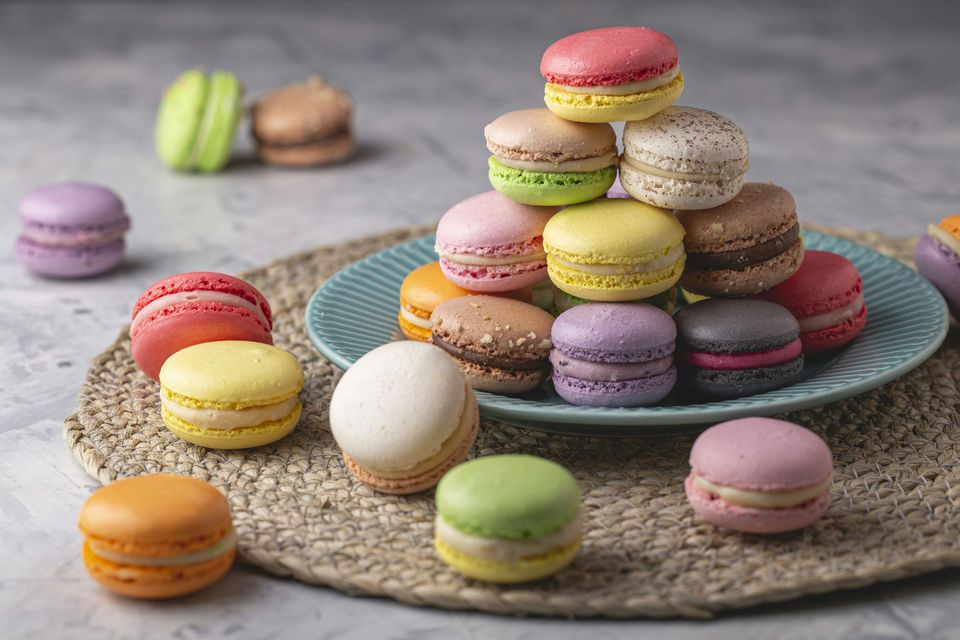 A pyramid of colourful macarons in pink, yellow, green, purple and chocolate shades on a plate, with more scattered across a woven placemat.