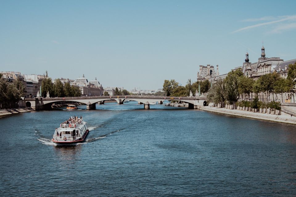 A sightseeing boat cruising the Seine on a clear day, passing beneath a stone bridge with tree-lined quays and Paris rooftops along the riverbank.