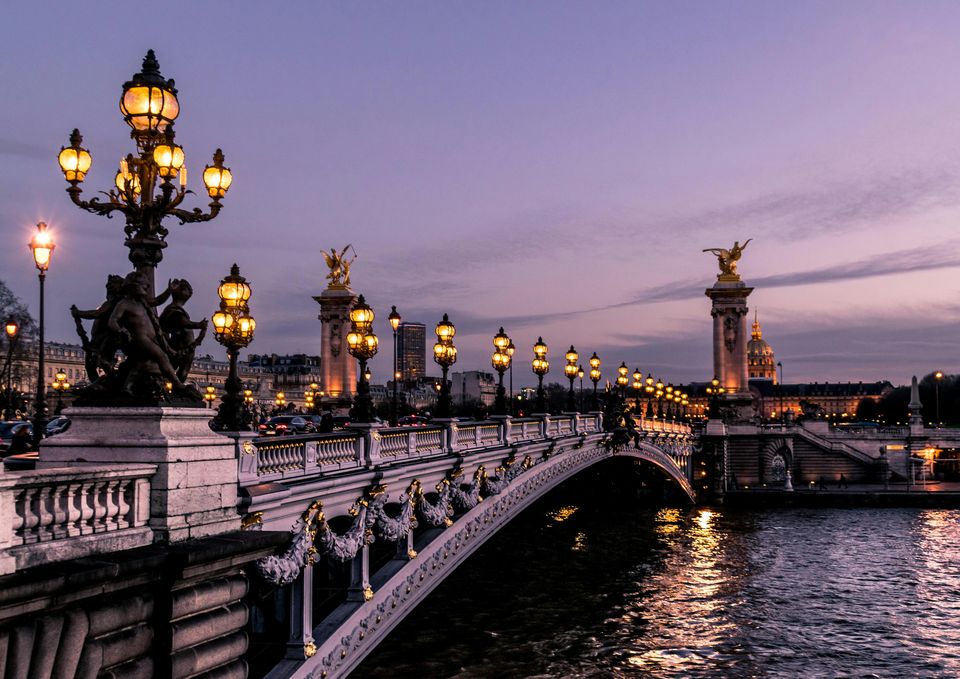 A dusk view along the Pont Alexandre III, its gilded lamps and statues glowing above the Seine with the dome of Les Invalides in the distance.