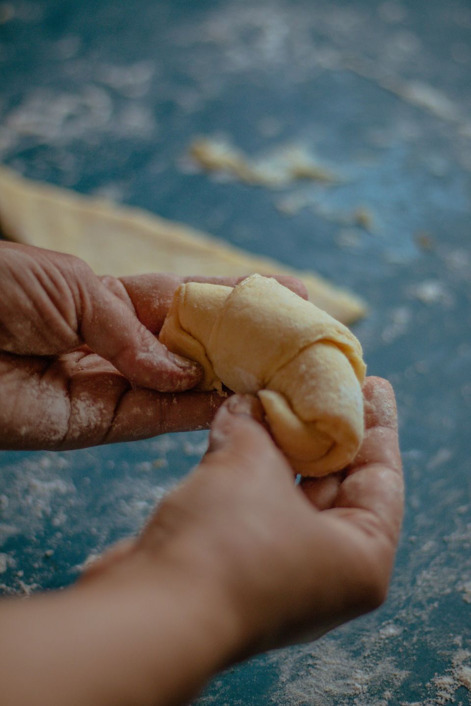 Two hands shaping a piece of croissant dough into its curved form on a flour-dusted work surface, with a sheet of rolled dough visible behind.